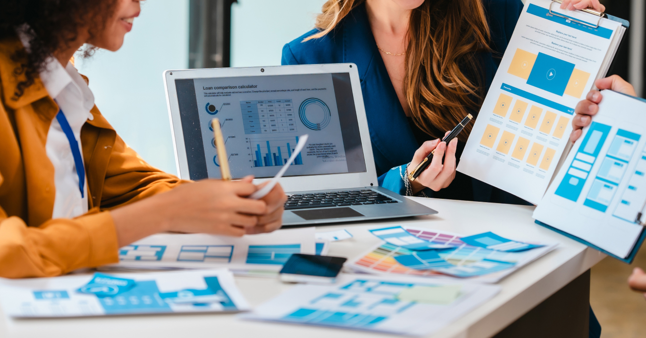 Imagem de pessoas trabalhando numam mesa branca, com um notebook e vários papeis de análise.